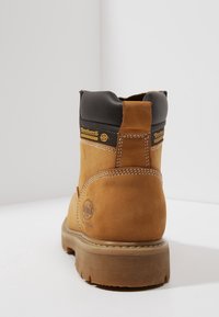 Tan læder arbejdsstøvle med sort polstret krave, mærke-patches, tykk gummisål og synlige sømme, set fra bag på hvid baggrund.