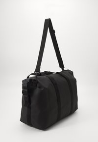 Sac de sport noir fabriqué dans un matériau durable ; doté d'une bandoulière, de deux poignées de transport et d'un design minimaliste sans logos visibles.
