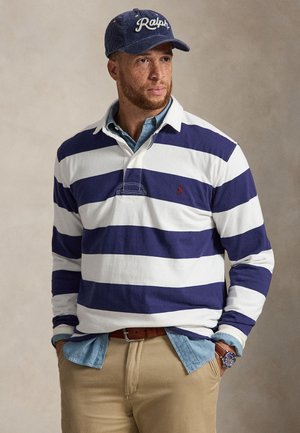 Mand iført en blå og hvidstribet rugbytrøje over en denimskjorte, khakibukser, en blå Ralph baseballkasket og et armbåndsur, stående med hænderne i lommerne.