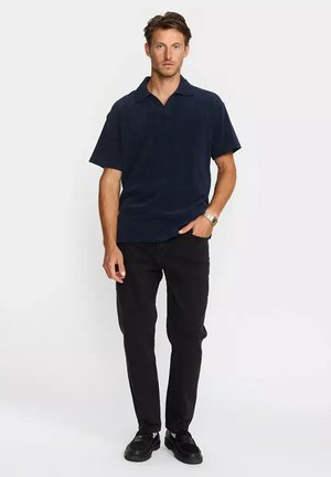 Homme debout portant une chemise bleu marine foncé à manches courtes avec col, un pantalon noir, des chaussures noires et une montre-bracelet au poignet gauche.