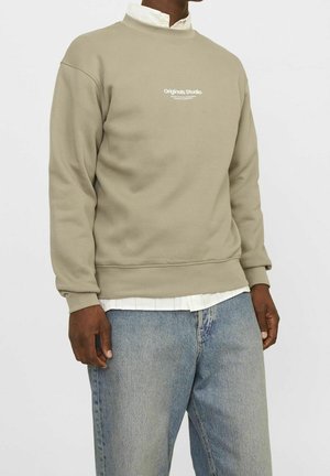 Person iført beige sweatshirt med teksten "Originals Studio", hvid skjorte med krave under og lyseblå jeans mod en ensfarvet baggrund.