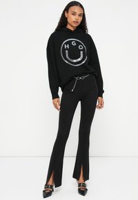 Zwarte hoodie met zilveren ronde logo, gecombineerd met zwarte uitlopende broek met zijsplitten. Model draagt puntige zwarte schoenen met banddetails.
