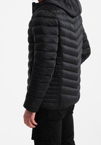 Sort isoleret jakke med hætte, med et quiltet design og vandrette striber. Materialet virker glat og puffy. Sorte bukser.