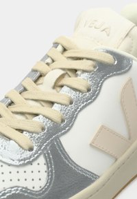 Close-up of beige-laced sneaker with silver, white, and light pink panels, showing perforated toe and stitched "V" logo on the side.