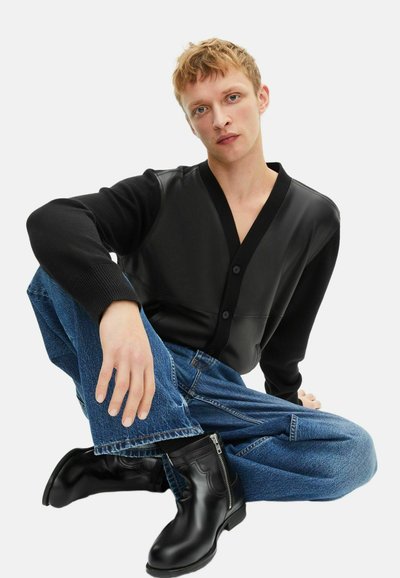 Ung mand sidder med korslagte ben iført en cardigan med sorte læderpaneler, blå jeans og sorte læder ankelstøvler med lynlås.