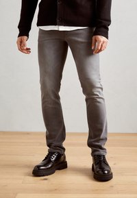 Man i grå slim-fit jeans, svarta läder snörskor, svart cardigan och vit skjorta står på ett trägolv.