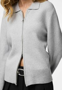 Cardigan lavorato a maglia grigio chiaro con scollo a collo, zip frontale e orlo leggermente corto, abbinato a una cintura nera con fibbia argentata.