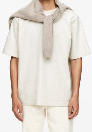 Homme portant une chemise beige clair à manches courtes avec un pull taupe drapé sur les épaules, associé à un pantalon blanc cassé.