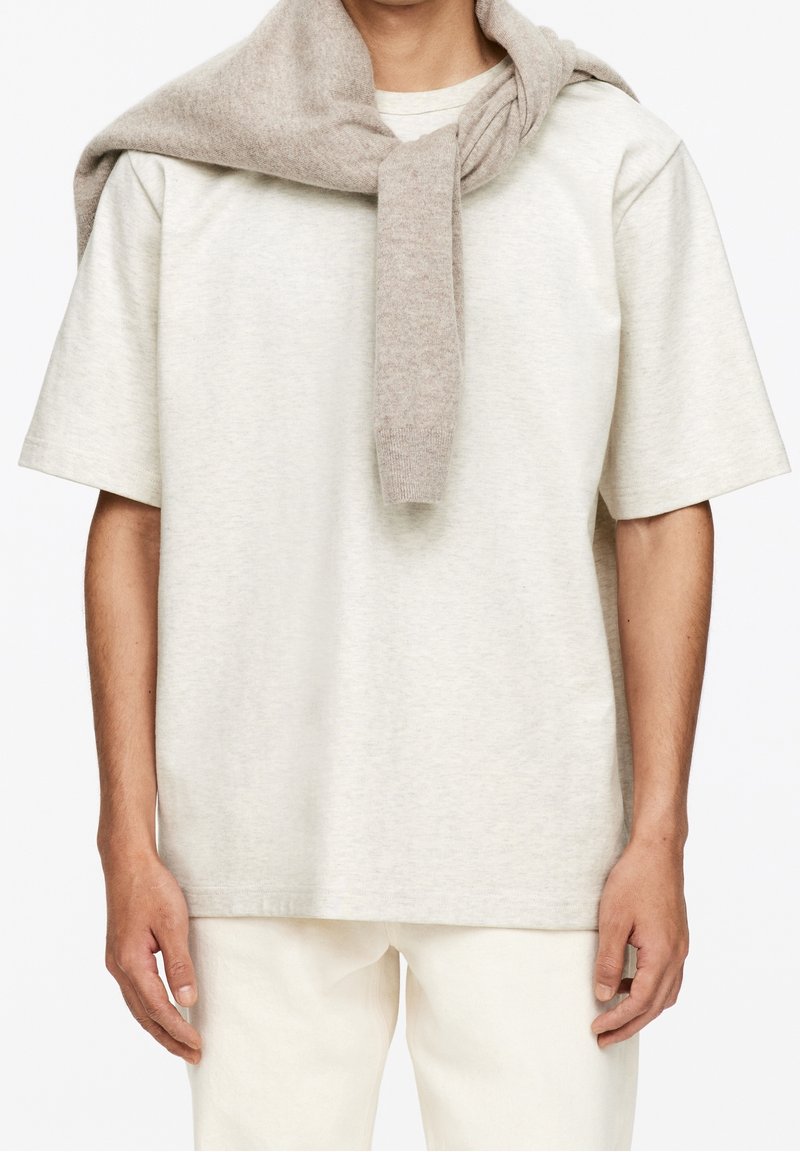 Homme portant une chemise beige clair à manches courtes avec un pull taupe drapé sur les épaules, associé à un pantalon blanc cassé.