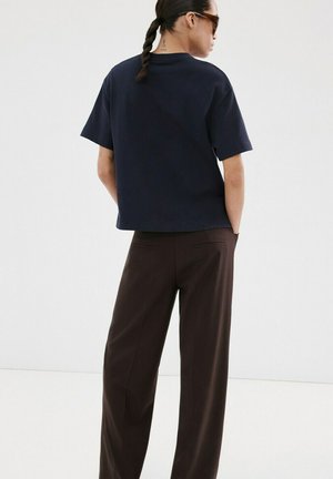 Frau mit geflochtenem Haar, die eine Sonnenbrille, ein marineblaues kurzärmeliges Hemd und dunkelbraune Hosen mit weitem Bein trägt und mit den Händen in den Taschen steht.