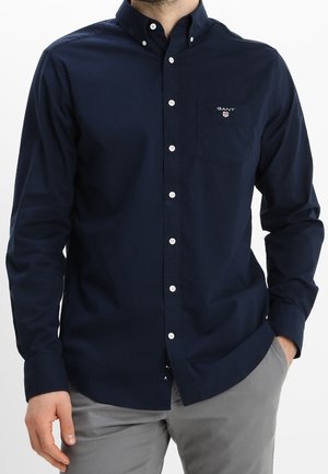 Homme portant une chemise bleu marine boutonnée avec une poche poitrine et le logo GANT, associée à un pantalon gris clair, debout avec une main dans la poche.
