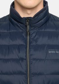 Marineblaue Steppjacke mit hohem Kragen, Reißverschluss vorne und horizontalem Steppmuster. Besitzt eine glänzende Oberfläche und dezente Branding-Elemente.