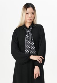 Young woman with long straight hair wears black dress with puffed sleeves and black scarf with white polka dots, standing against white background.