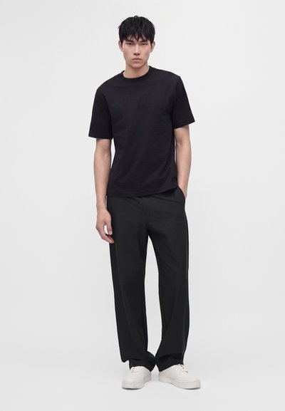 Jeune homme debout face à la caméra, portant un t-shirt noir à manches courtes, un pantalon noir et des baskets blanches sur un fond clair uni.