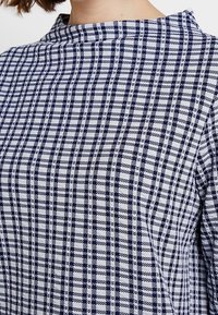 Marinblå och vit rutigt topp med hög krage, med en strukturerad tyg och växlande vertikala och horisontella ränder i ett rutmönster.