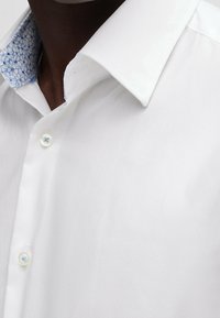 Witte shirt met een gladde textuur, contrasterende lichtblauwe kraag met bloemenpatroon, en duidelijke knoopsluiting.