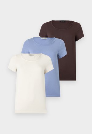 Kvinne iført mørkebrun kortermet topp, beige vide cargobukser og beige flate sko, stående mot en ensfarget hvit bakgrunn.