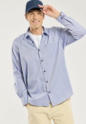 Homme souriant, portant une chemise bleue à boutons, un t-shirt blanc, un pantalon beige et une casquette bleu marine, touchant la visière de la casquette avec sa main droite.