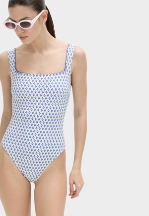 Femme aux cheveux bruns attachés en queue de cheval, portant des lunettes de soleil blanches et un maillot de bain une pièce blanc avec de petits motifs losanges bleus.