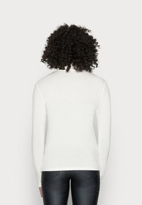 Person mit lockigem Haar, der ein enges, weißes Langarmoberteil und schwarze Lederhosen trägt und mit dem Rücken zu einem schlichten Hintergrund steht.