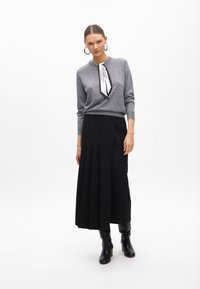 Szary sweter z dzianiny o okrągłym dekolcie i czarno-białym detalem w postaci wstążki, zestawiony z czarną plisowaną maxispódnicą i czarnymi kozakami do kolan.