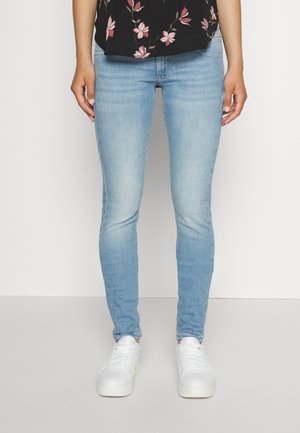 Person iført lyseblå skinny jeans, hvide sneakers og en sort blomstret top, stående mod en hvid baggrund.