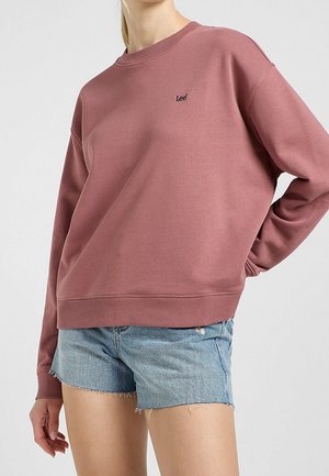 Kvinde iført en støvet rosa Lee-sweatshirt og lyseblå frynsede denimshorts, stående med den ene hånd på hoften mod en ensfarvet baggrund.