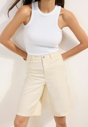 Žena v bielej bezrukávovej rebrovanej blúzke a krémových krátkych nohaviciach s vysokým pásom do kolien, stojaca s rukami na bokoch pred jednofarebným pozadím.
