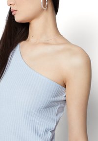 Světle modrý žebrovaný top na ramínka s asymetrickým designem na jednom rameni. Modelka má na sobě stříbrné kroužkové náušnice a jemný stříbrný náhrdelník.