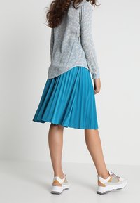 Gonna plissettata teal con un design fluido, abbinata a una maglietta a maniche lunghe azzurro chiaro con un motivo delicato e sneakers bianche con dettagli.