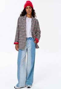 Žena v červené čepici, kostkovaném saku s červenými manžetami, bílém tričku, širokých dvoubarevných modrých džínách a bílých teniskách, stojící s jednou rukou v kapse.