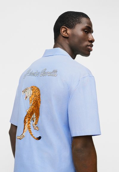 Άνδρας που φοράει μπλε πουκάμισο με κοντά μανίκια και ρίγες, με κεντημένη βρυχώμενη τίγρη και το κείμενο «Roberto Cavalli» στην πλάτη, κοιτάζει προς τα δεξιά του.