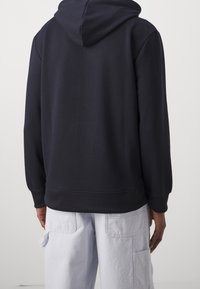 Marinblå hoodie med avslappnad passform, ribbade mudd och nederkant. Har en stor huva och slät textur, bärs med ljusfärgade cargobyxor.