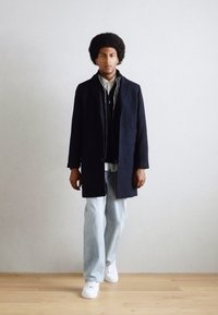 Abrigo negro de corte recto, llevado sobre un chaleco de punto azul y una camisa blanca. Unos vaqueros azul claro y unas zapatillas blancas completan el conjunto.