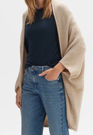 Femme portant un cardigan beige en tricot, une chemise bleu marine et un jean bleu, une main dans la poche avant.