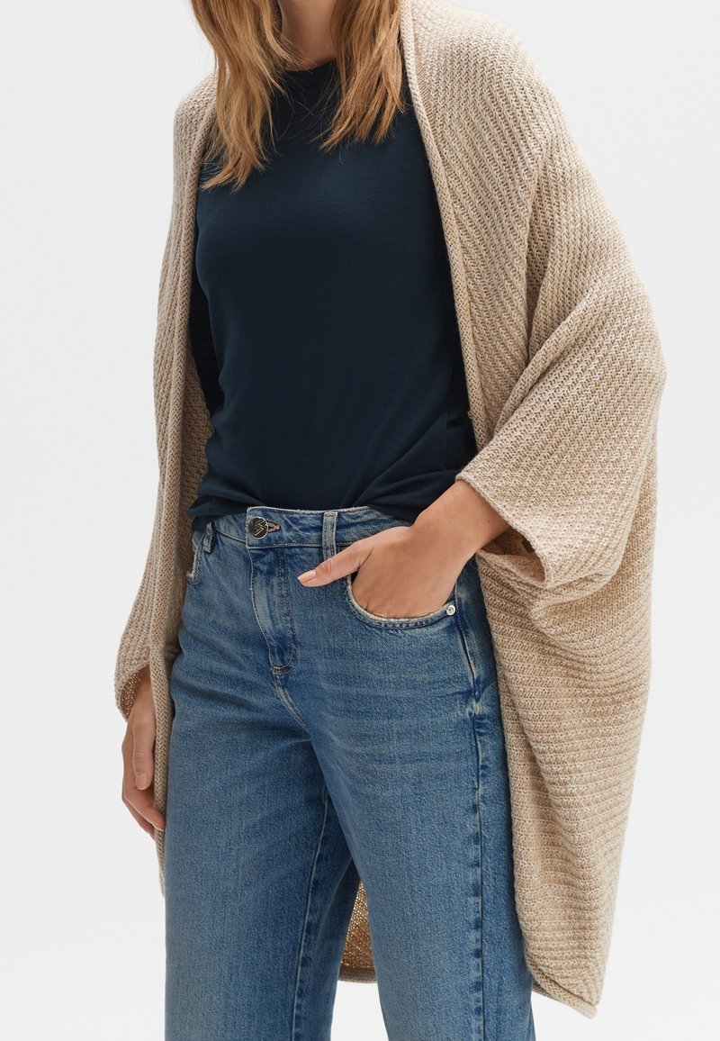 Femme portant un cardigan beige en tricot, une chemise bleu marine et un jean bleu, une main dans la poche avant.