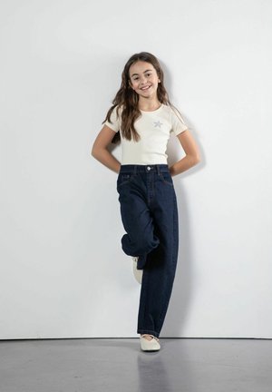 Uśmiechnięta dziewczyna o długich brązowych włosach, ubrana w białą koszulkę z motywem gwiazdy, ciemnoniebieskie dżinsy i białe buty, stojąca na tle białej ściany.