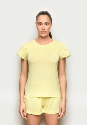 Femme debout sur fond blanc portant un haut à manches courtes jaune clair avec des détails en dentelle et un short assorti, face à la caméra avec une expression neutre.