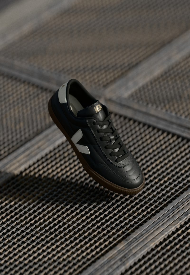 Sapatilha de couro preto com detalhes cinzentos, atacadores e sola de borracha. Apresenta um logótipo branco proeminente e um design de parte superior texturizada.