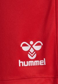 Czerwony poliesterowy t-shirt sportowy z białym logo o abstrakcyjnym wzorze oraz słowem "hummel" napisanym wyraźną czcionką. Gładka faktura.