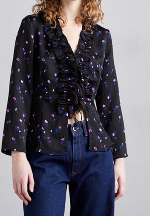 Vrouw draagt een zwarte blouse met lange mouwen met paarse bloemenprint en ruches aan de voorkant, gecombineerd met blauwe high-waist jeans tegen een effen achtergrond.