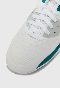 Λευκό αθλητικό παπούτσι με επάνω μέρος από υφασμάτινο πλέγμα, γκρι λεπτομέρειες και τυρκουάζ εξωτερική σόλα. Σχεδιασμένο με επίπεδα λευκά κορδόνια και στρογγυλερό δάχτυλο.