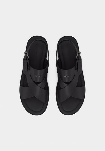 Ζευγάρι μαύρα δερμάτινα σανδάλια με σταυρωτά λουριά, ρυθμιζόμενες αγκράφες στον αστράγαλο και λογότυπο "BALLY" στα πάνω λουριά, τοποθετημένα σε ανοιχτό φόντο.