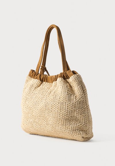 Μπεζ υφασμάτινη τσάντα ώμου με καφέ δερμάτινες λαβές και ρυτιδωμένη κορυφή, παρουσιασμένη μπροστά σε απλό φωτεινό φόντο.