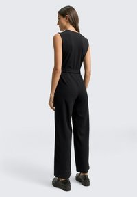 Une femme aux longs cheveux bruns porte une combinaison sans manches noire et des mocassins noirs, debout et tournée vers le mur contre un fond gris uni.