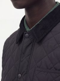 Gros plan d'une veste matelassée foncée avec un col en velours côtelé noir et des boutons-pression portée sur une chemise verte.