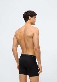 Man met donker haar die zwarte Calvin Klein boxershort draagt, staat bloot bovenlijf, met zijn gezicht naar achteren tegen een neutrale grijze achtergrond.
