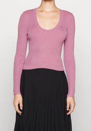 Femme portant un pull rose ajusté à manches longues en tricot côtelé avec un col en U et une jupe plissée noire taille haute.