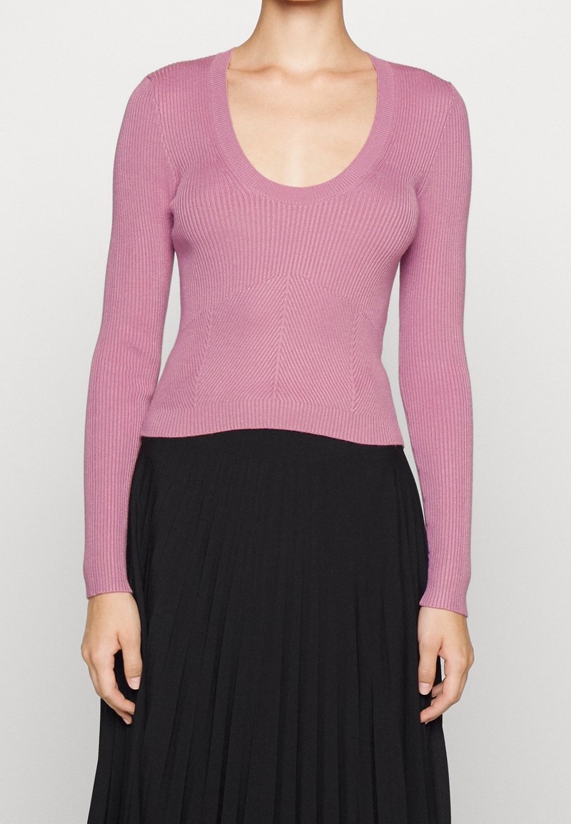 Femme portant un pull rose ajusté à manches longues en tricot côtelé avec un col en U et une jupe plissée noire taille haute.