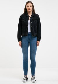 Fekete farmerdzseki elülső zsebekkel és gombos zárással, kék skinny farmernadrággal és fekete sneaker cipővel párosítva. Sima textúra, klasszikus fazon.